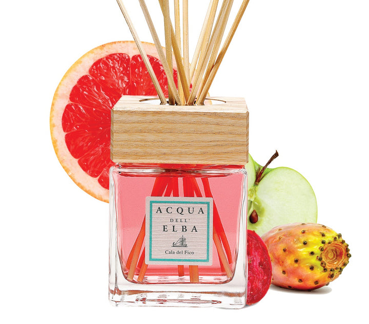 Acqua di Elba diffuser bottle with pink liquid, a light wood cap, and natural reed sticks, styled with grapefruit, strawberry, and green apple slices on a white background.